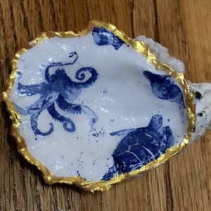 May include: A white oyster shell with a gold rim, painted with a blue octopus and a blue sea turtle.