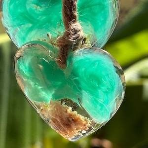 May include: Two teal blue resin hearts with a brown twine string. The hearts are filled with a swirling pattern of teal blue resin and have a small piece of dried flower inside.