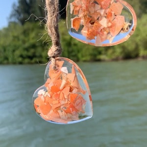 May include: Two clear heart-shaped ornaments filled with small pieces of orange and white material. The ornaments are hanging from a brown twine string.