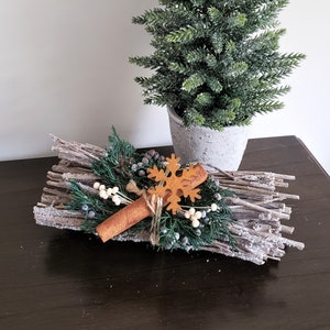 May include: A rustic winter centerpiece featuring a cinnamon stick, a felt snowflake, and sprigs of greenery on a bed of white-washed twigs.