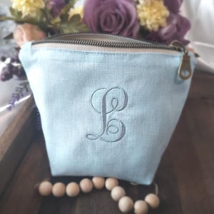 May include: A light blue zippered pouch with a gray embroidered letter "B" on the front. The pouch has a beige zipper and a bronze-colored zipper pull. A wooden bead strand is in the foreground, and flowers are in the background.