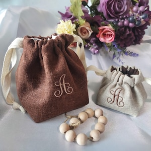 May include: Two drawstring pouches, one brown and one beige, each embroidered with the letter "A". The pouches have cream-colored drawstrings and are set against a backdrop of flowers and a wooden bead bracelet.