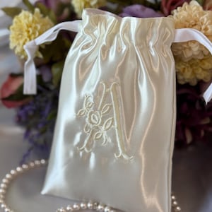 May include: Ivory satin drawstring bag with an embroidered floral initial "A". The bag is gathered at the top with white satin ribbons. A pearl necklace is in the foreground, and a bouquet of flowers is in the background.