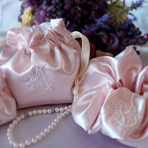 May include: A set of pale pink satin drawstring pouches and a matching scrunchie, each featuring white embroidered monogram letters. The items are accompanied by a strand of white faux pearls, showcasing a smooth, reflective fabric finish.