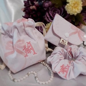 May include: A set of matching accessories in a soft pink hue, including a drawstring bag, a small wallet, and a scrunchie. Each item features a delicate bow pattern and the embroidered letter "A". A pearl necklace is also present.