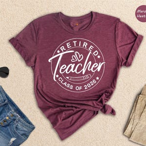 a maroon t-shirt with a white graphic design that says "Retired Teacher CLASS OF 2026" and is displayed on a beige background.