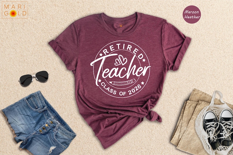 a maroon t-shirt with a white graphic design that says "Retired Teacher CLASS OF 2026" and is displayed on a beige background.