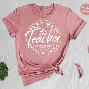 a pink t-shirt with a white design that says "Retired Teacher Class of 2026" and a rose next to it. There are also some feathers scattered around the t-shirt and a small plant.