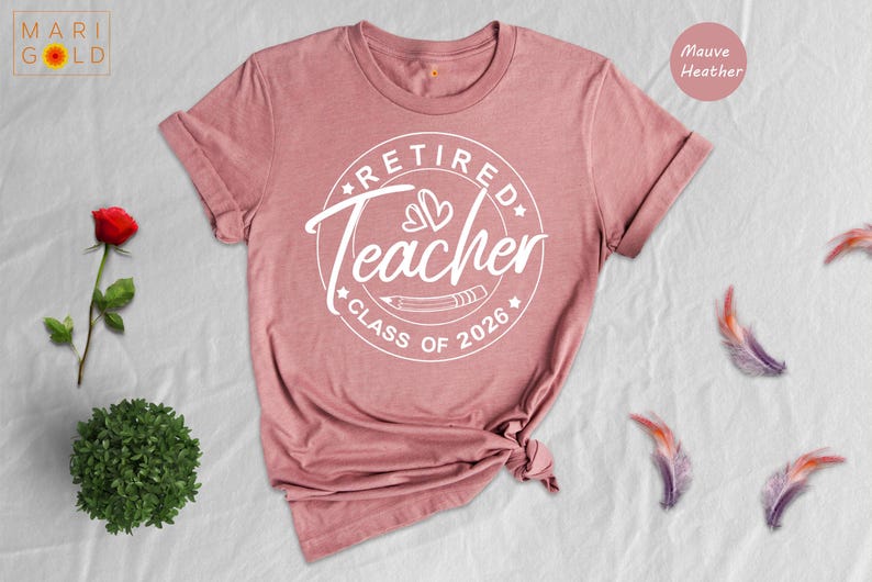 a pink t-shirt with a white design that says "Retired Teacher Class of 2026" and a rose next to it. There are also some feathers scattered around the t-shirt and a small plant.