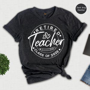 a grey t-shirt with a white graphic design that says "Retired Teacher Class of 2026" printed on it. The t-shirt is displayed on a white background, along with a pair of denim shorts, a notebook, a pen, and a plant.