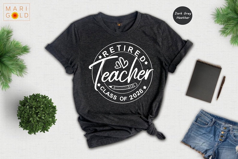 a grey t-shirt with a white graphic design that says "Retired Teacher Class of 2026" printed on it. The t-shirt is displayed on a white background, along with a pair of denim shorts, a notebook, a pen, and a plant.