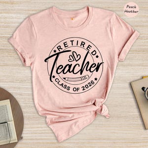 a pink t-shirt with a graphic design that says "Retired Teacher CLASS OF 2026" printed on it. The t-shirt is displayed on a wooden surface, along with a cup, a notebook, and a pen.