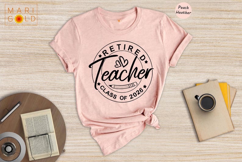 a pink t-shirt with a graphic design that says "Retired Teacher CLASS OF 2026" printed on it. The t-shirt is displayed on a wooden surface, along with a cup, a notebook, and a pen.