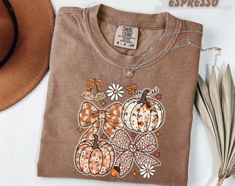 May include: Espresso-colored t-shirt with a crew neck and a Comfort Colors tag. The shirt features a fall-themed graphic with pumpkins, bows, and floral accents. A silver necklace is draped across the shirt.