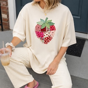 May include: A cream-coloured t-shirt with a large, patchwork strawberry motif. The strawberry design incorporates various fabric patterns, such as red gingham, floral, and heart prints. The shirt has short sleeves and a relaxed fit, ideal for everyday wear.