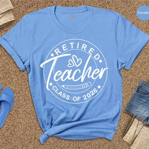 A blue t-shirt with a white design that says "Retired Teacher CLASS OF 2026" is displayed on a beige surface, along with a pair of blue sandals and a pair of denim shorts.