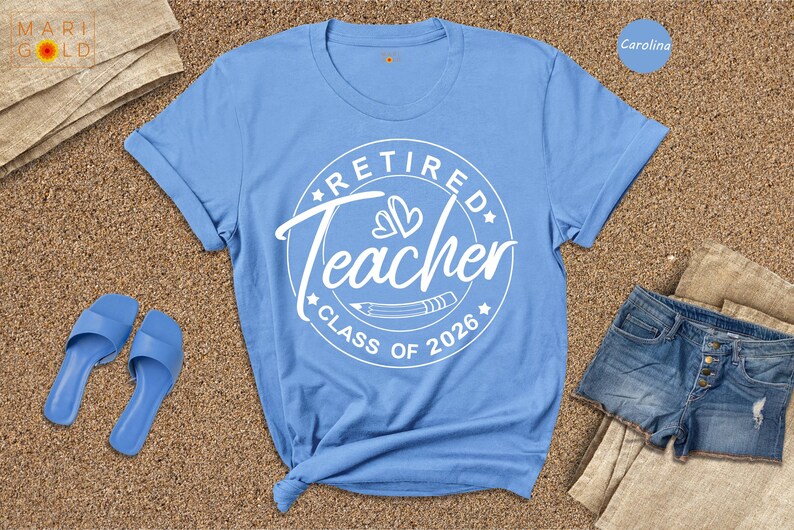 A blue t-shirt with a white design that says "Retired Teacher CLASS OF 2026" is displayed on a beige surface, along with a pair of blue sandals and a pair of denim shorts.