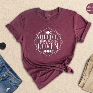 May include: A maroon heather t-shirt with the text 'Support your local coven' in a white circle with a geometric design and moon phases. The t-shirt is folded and displayed on a light beige surface.