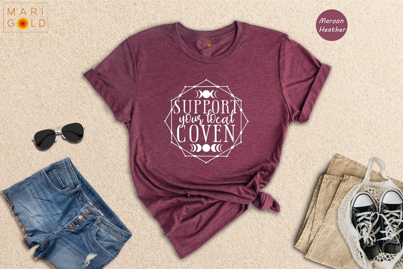 May include: A maroon heather t-shirt with the text 'Support your local coven' in a white circle with a geometric design and moon phases. The t-shirt is folded and displayed on a light beige surface.