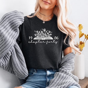 May include: Dark gray t-shirt with a white floral design above the text "1986 chapter forty." The design features an open book with flowers and foliage. The shirt is a crew neck and is worn by a person.