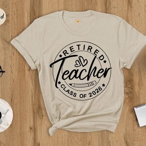 a beige t-shirt with a graphic design that says "Retired Teacher CLASS OF 2026" printed on it, along with a pencil and a cup of coffee. The t-shirt is placed on a wooden surface, and there are other items such as a pair of glasses, a notebook, and a potted plant nearby.