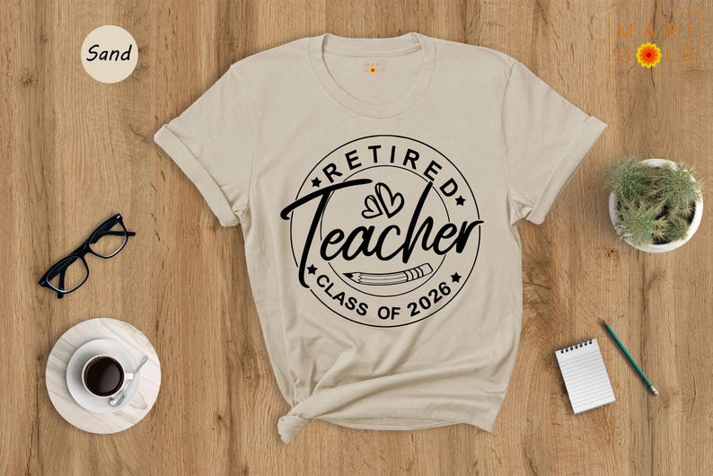 a beige t-shirt with a graphic design that says "Retired Teacher CLASS OF 2026" printed on it, along with a pencil and a cup of coffee. The t-shirt is placed on a wooden surface, and there are other items such as a pair of glasses, a notebook, and a potted plant nearby.