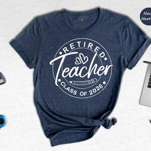 a navy blue t-shirt with a white graphic design that says "Retired Teacher CLASS OF 2026" printed on it. The t-shirt is laid out on a white surface, along with a laptop, a camera, and some headphones.