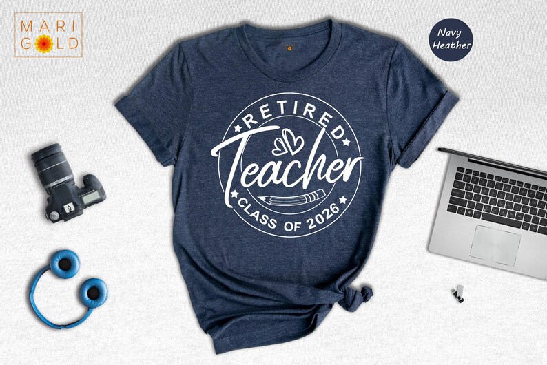 a navy blue t-shirt with a white graphic design that says "Retired Teacher CLASS OF 2026" printed on it. The t-shirt is laid out on a white surface, along with a laptop, a camera, and some headphones.