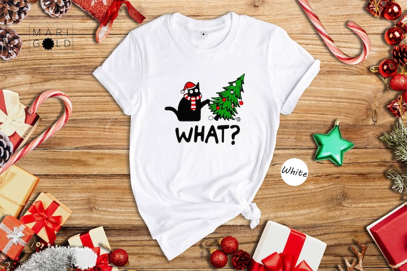 May include: White t-shirt with a black cat wearing a Santa hat and scarf pushing over a green Christmas tree with ornaments. The text 'WHAT?' is printed below the image.