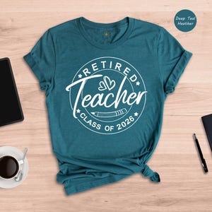 a teal t-shirt with a white design that says "Retired Teacher CLASS OF 2026" on it, along with a black notebook, a tablet, a cup of coffee, and a pen.
