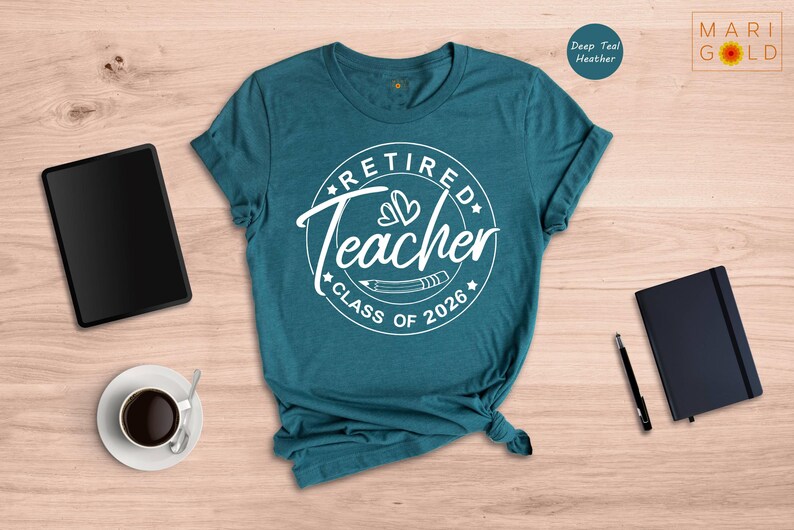 a teal t-shirt with a white design that says "Retired Teacher CLASS OF 2026" on it, along with a black notebook, a tablet, a cup of coffee, and a pen.