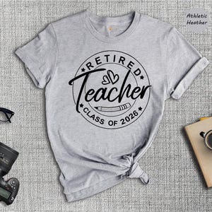 a gray t-shirt with a graphic design that says "Retired Teacher Class of 2026", along with a pair of glasses, a camera, and a book.