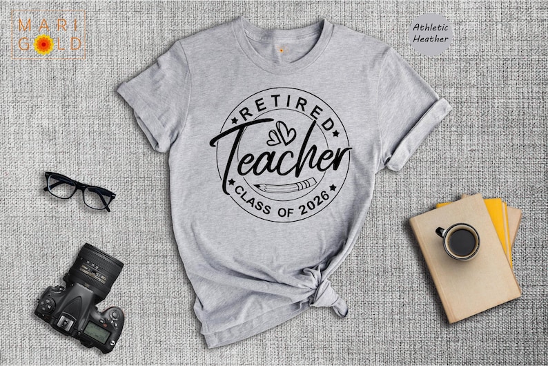 a gray t-shirt with a graphic design that says "Retired Teacher Class of 2026", along with a pair of glasses, a camera, and a book.