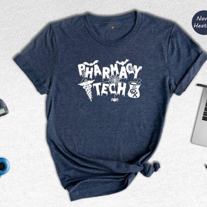 May include: A navy heather t-shirt with the text 'Pharmacy Tech' in white. The text is surrounded by Halloween-themed graphics, including bats, a spider web, a cauldron, and a caduceus.