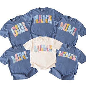 May include: A collection of blue and cream-colored t-shirts. Each shirt features a different name, such as "GIGI", "NANA", "MIMI", "MAMA", and "MEMAW", in a patchwork design. The shirts are casual and comfortable.