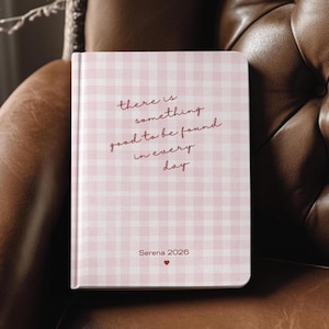 Puede incluir: Un cuaderno con un patrón de cuadros vichy rosa y blanco. La portada presenta el texto manuscrito "there is something good to be found in every day". También incluye el nombre "Serena 2026" y un símbolo de corazón.