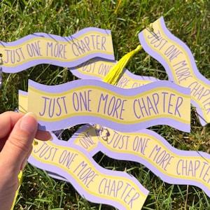 May include: A set of wavy, light purple and yellow bookmarks featuring the text "JUST ONE MORE CHAPTER" in a dark purple font. Each bookmark is attached to a bright yellow tassel, creating a collection of identical reading accessories.