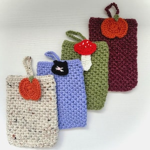 May include: Four rectangular crochet pouches with hanging loops. The first is speckled beige with an orange pumpkin, the second is light purple with a black cat face, the third is olive green with a red mushroom, and the fourth is dark maroon with an orange pumpkin.