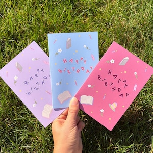 May include: Three rectangular greeting cards in light purple, light blue, and pink. Each card features the text "HAPPY BIRTHDAY" in a playful font, surrounded by small illustrations of open books, closed books, white flowers, and tiny candles.