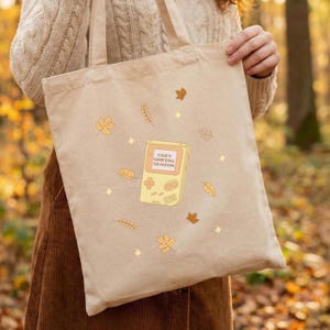 Könnte beinhalten: Eine beigefarbene Tragetasche aus Segeltuch mit einer zentralen Grafik einer gelben tragbaren Spielkonsole, auf deren Bildschirm der Text COZY GAMING SEASON steht, umgeben von verstreuten orangefarbenen und gelben Herbstblättern und kleinen vierzackigen Sternen.