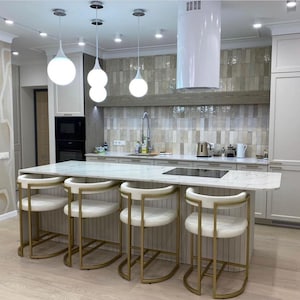May include: A modern kitchen island with a white marble worktop and four gold bar stools. The island has a built-in electric hob and a white range hood.