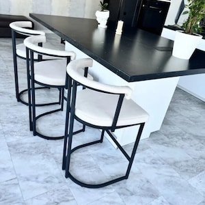 May include: Three black metal bar stools with white cushioned seats. The stools are arranged in a row in front of a black countertop.
