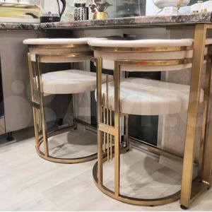 Könnte beinhalten: Zwei goldene Barhocker mit weißen gepolsterten Sitzen. Die Hocker haben ein geometrisches Design und befinden sich in einer Küche.