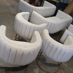 Op de afbeelding: Een set van zes witte, gebogen stoelen met verticale kanaalstoffering. De stoelen hebben een afgeronde rugleuning en zitting, ondersteund door goudkleurige metalen poten. De stoelen zijn opgesteld in een werkplaats.