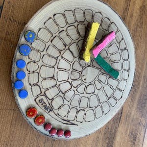May include: A circular wooden board with a wood-burned spiral path and the word START. The board features small colourful circular game pieces in blue, yellow, red, and maroon, along with three rectangular wooden markers painted yellow, pink, and green.