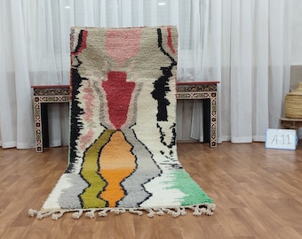 May include: A rectangular, colorful rug with a geometric design. The rug features a central pattern with red, orange, and yellow hues, bordered by black, white, and green sections. Tassels adorn the bottom edge. The rug is displayed on a wooden floor.