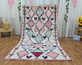May include: A white, pink, and blue Moroccan rug with a diamond pattern. The rug has a fringe border and is made of wool.