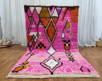 Puede incluir: Una alfombra marroquí rosa y naranja con un patrón geométrico. La alfombra tiene un borde de flecos y está hecha de lana.