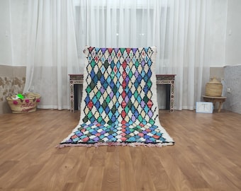 May include: A colorful Moroccan rug with a diamond pattern in shades of blue, green, red, and white. The rug is laid out on a wooden floor.