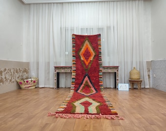 May include: A long, red, and green Moroccan rug with a diamond pattern. The rug has a fringe on both ends and is laid out on a wooden floor. The rug is in front of a white curtain and a small table with a vase on it.